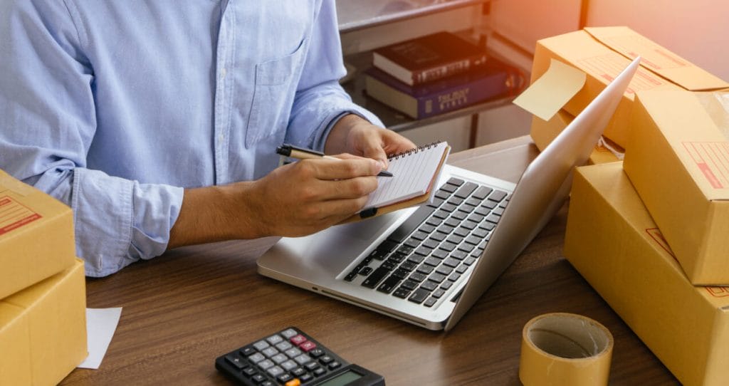 A man holding a pen and a mini notebook, sitting at a desk with a laptop computer, boxes, and a calculator | eCommerce marketing