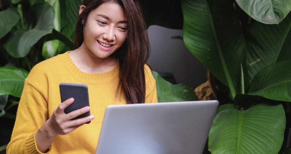 A girl wearing a yellow sweater is smiling, holding a phone, and looking at a laptop computer | Upgrade to Wordfence Premium